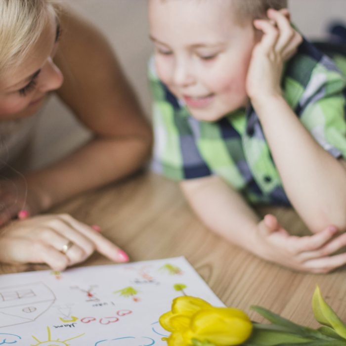 Kobieta o blond włosach i mały chłopiec leżą na podłodze, wspólnie oglądając dziecięcy rysunek. Kobieta wskazuje palcem na detale obrazka, a obok nich leży żółty tulipan.
